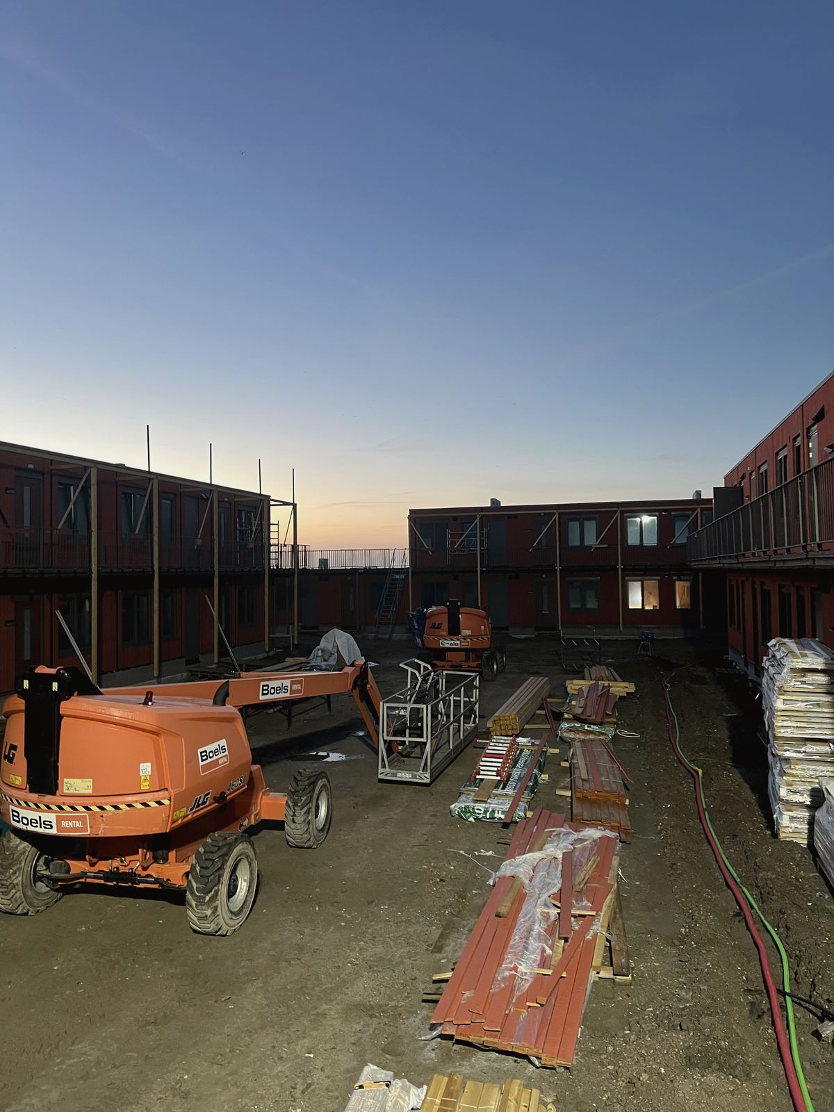 Bouwplaats van het nieuwe AZC in Opmeer bij zonsondergang, met hoogwerkers langs de houten buitengevels.